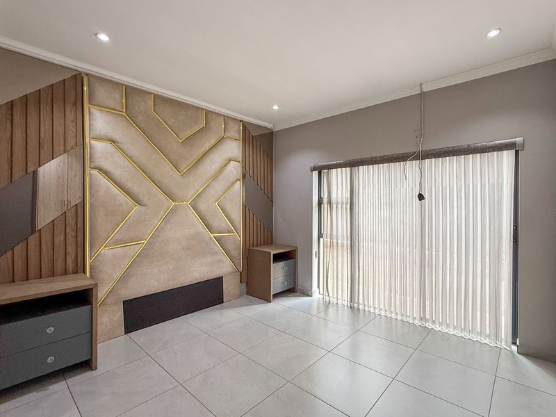 A modern bedroom featuring a decorative upholstered headboard wall, tiled flooring, and a sliding glass door with vertical blinds.