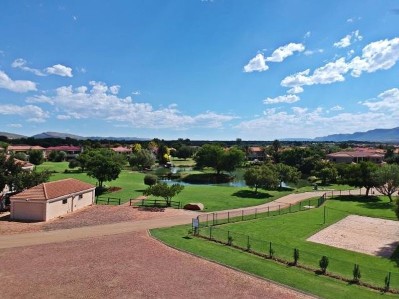 Aerial view of a residential estate featuring landscaped green spaces, a central lake, and recreational facilities under a clear blue sky.