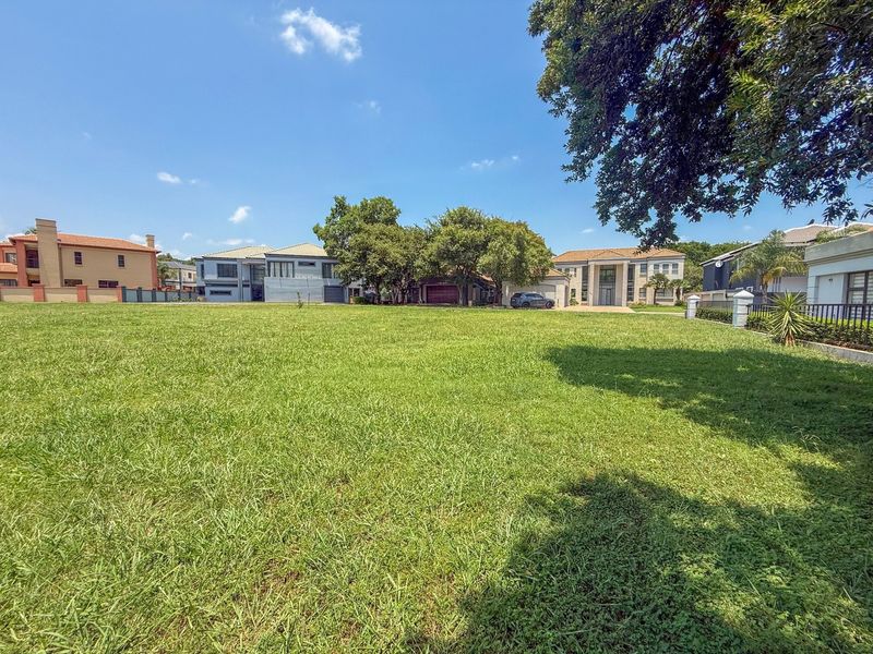 A spacious, level vacant residential plot featuring a well-maintained grass lawn, established trees, and perimeter fencing in a developed neighborhood.