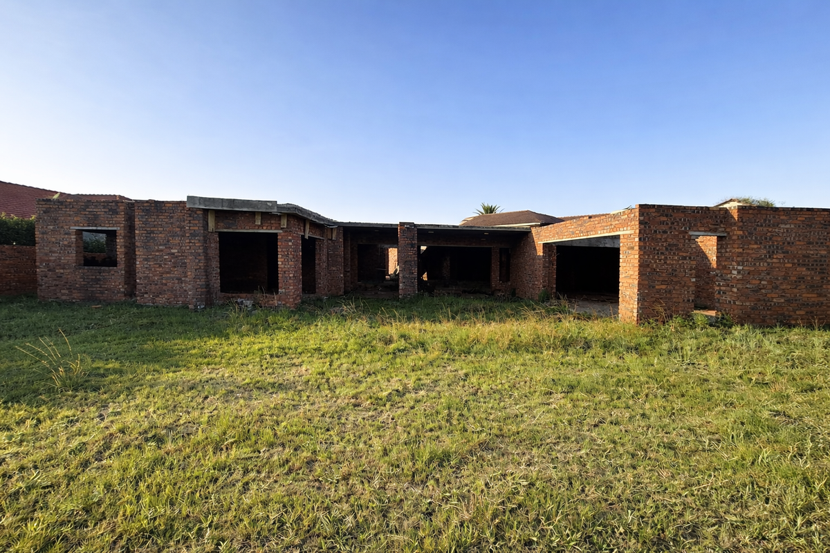An unfinished residential property structure showing exposed brickwork and structural openings, set on a large grassed plot.