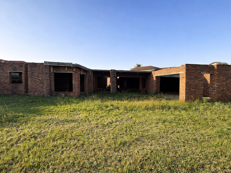 An unfinished residential property structure showing exposed brickwork and structural openings, set on a large grassed plot.
