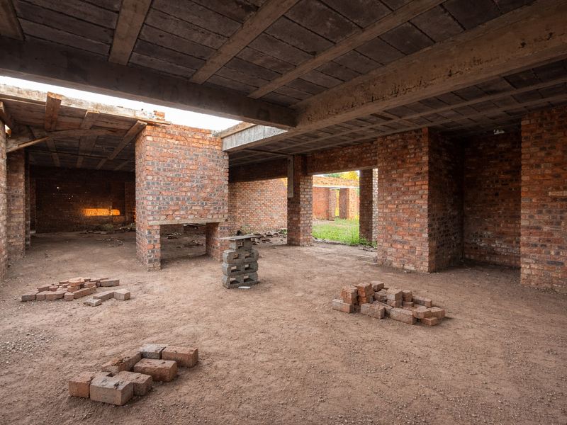 An unfinished construction site featuring exposed brick walls, structural brick pillars, and a concrete slab ceiling with a dirt floor.