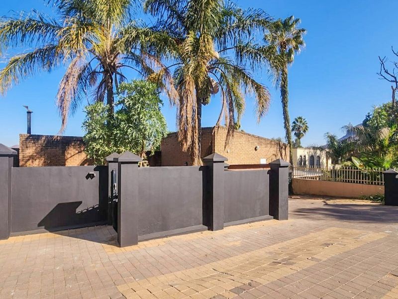 Front exterior view featuring a dark painted brick boundary wall, pedestrian gate, and paved driveway area with established palm trees.