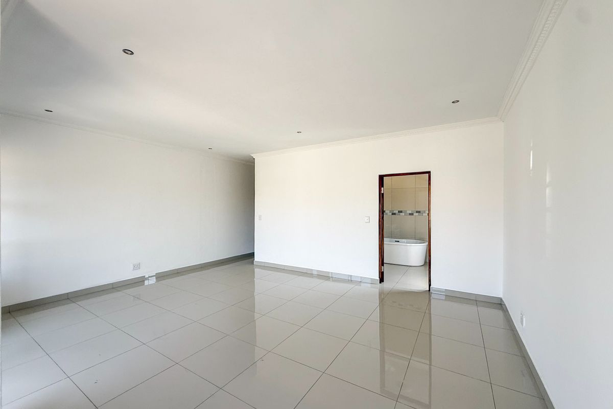 A spacious, empty bedroom featuring neutral tiled flooring, recessed lighting, and coved ceilings, with direct access to an en-suite bathroom.