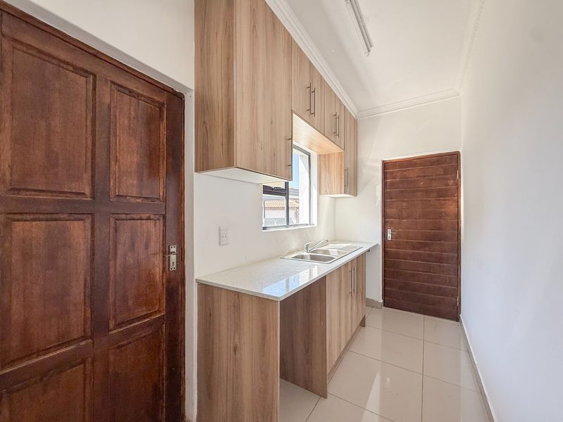A clean and functional scullery featuring modern wood-finish cabinetry, stone countertops, a stainless steel sink, and tiled flooring.