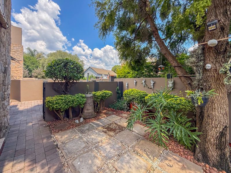 A well-maintained private garden area featuring paved patio sections, mature trees, and established shrubbery enclosed by a solid boundary wall.