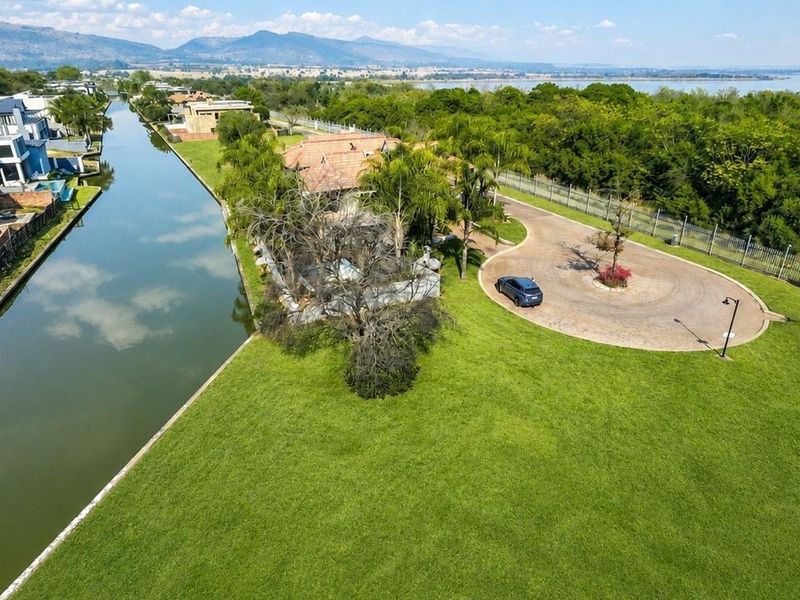 Aerial view of a residential vacant land plot situated along a canal, featuring a paved driveway and established landscaping with scenic water and mountain views.