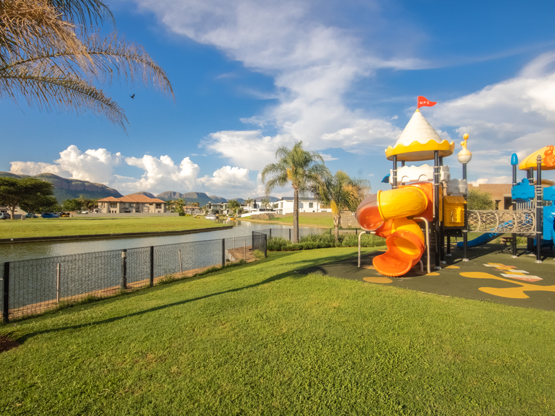 A well-maintained communal garden area featuring a playground, manicured lawn, and scenic water views within a residential estate.