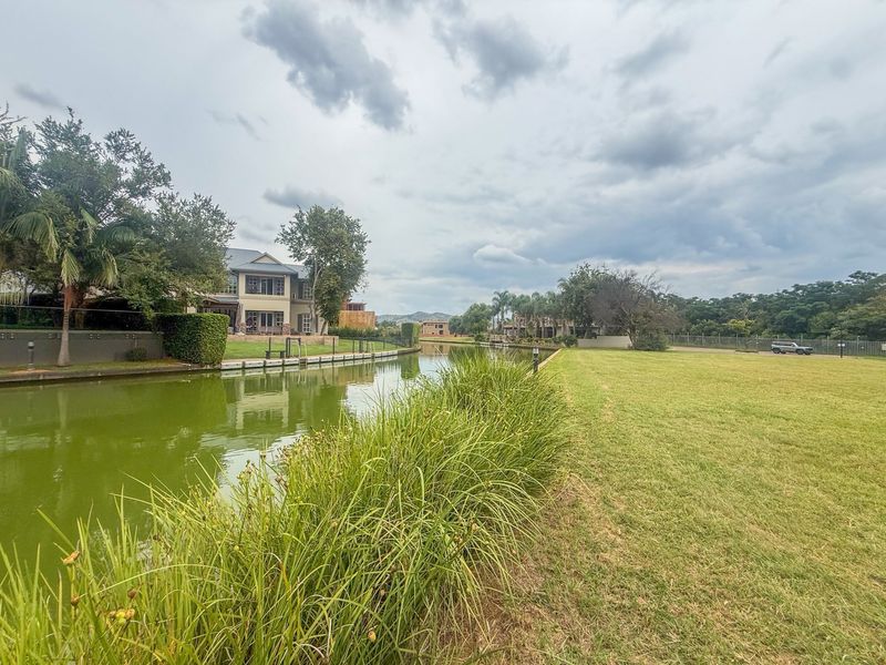 A scenic view of the waterfront area featuring a well-maintained lawn, natural vegetation, and surrounding residential properties.