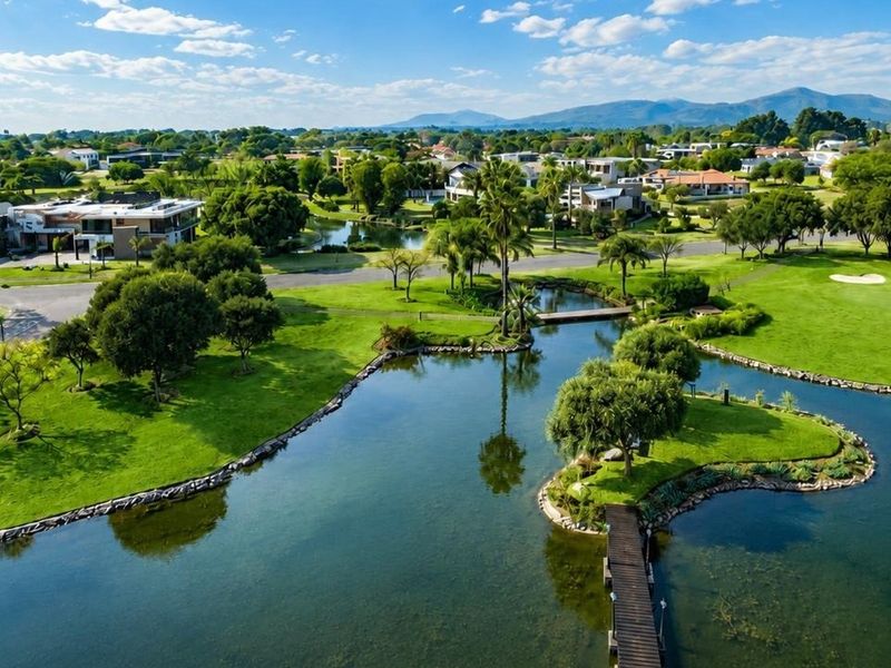 An aerial view of a residential estate featuring a central lake, landscaped green spaces, and a pedestrian bridge within a golf course community.