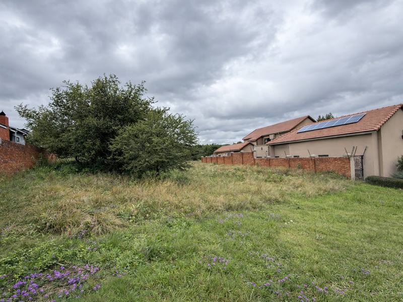 A vacant residential plot featuring established grass, a brick boundary wall with electric fencing, and a view of adjacent properties with solar panels.