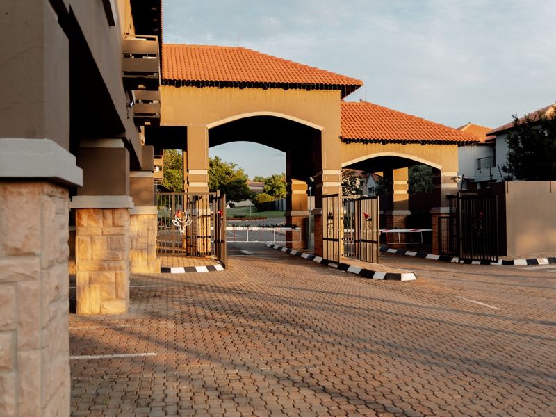A secure residential estate entrance featuring a paved driveway, stone-clad pillars, and automated boom gates under a tiled roof structure.
