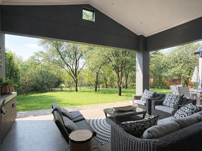 A spacious covered patio featuring a vaulted ceiling, recessed lighting, and a built-in braai, overlooking a lush green garden.