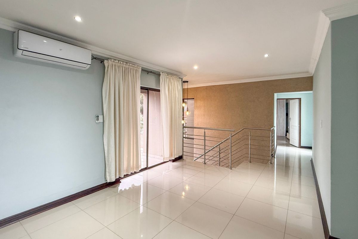 Spacious, light-filled living area featuring tiled flooring, a modern stainless steel staircase railing, and a wall-mounted split-system air conditioner.