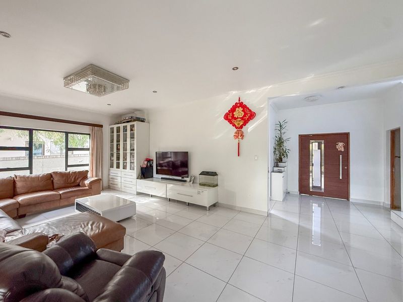 Spacious, light-filled living room featuring large-format white floor tiles, recessed lighting, a decorative chandelier, and a modern solid wood entrance door.