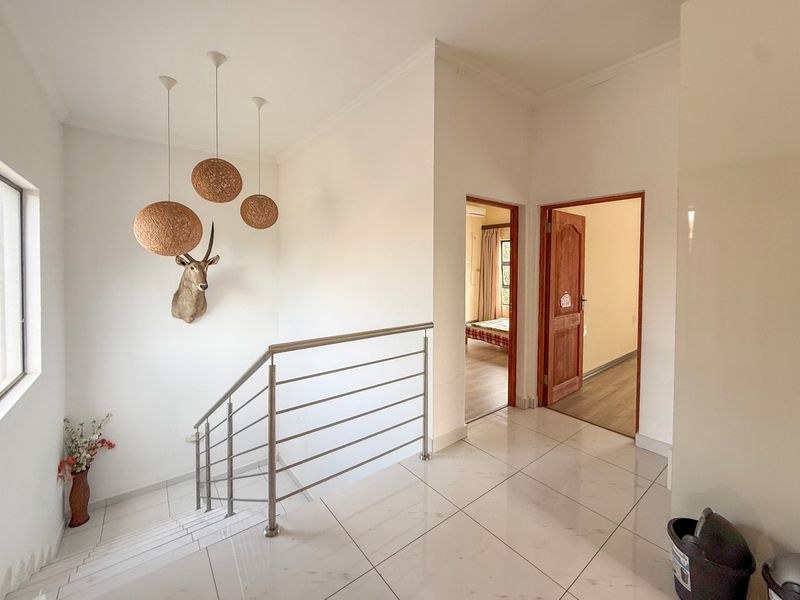 A bright, modern hallway featuring tiled flooring, a stainless steel staircase balustrade, and access to adjacent bedrooms through wooden doors.