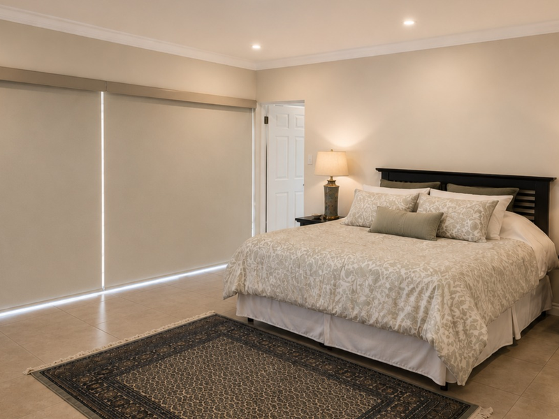 A spacious, well-maintained bedroom featuring neutral tiled flooring, recessed downlighting, and large roller blinds for privacy.