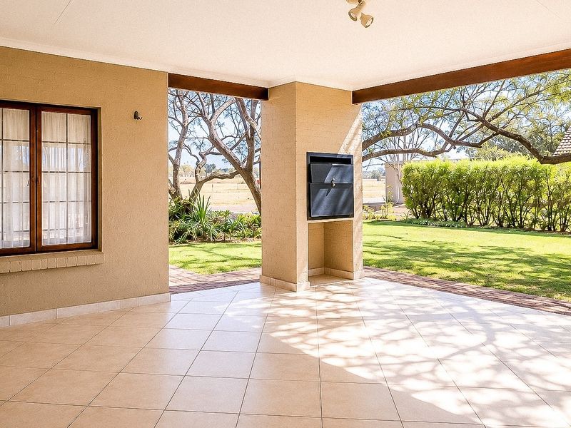Spacious covered patio featuring tiled flooring and a built-in braai, overlooking a well-maintained garden area.