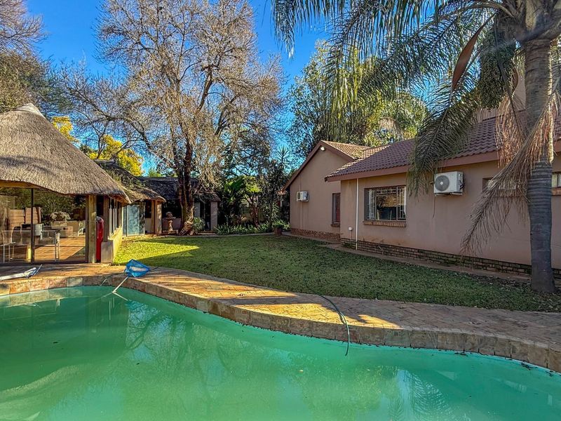 Outdoor pool area featuring a swimming pool, paved surround, and a thatched lapa, set within a garden space adjacent to the main house.