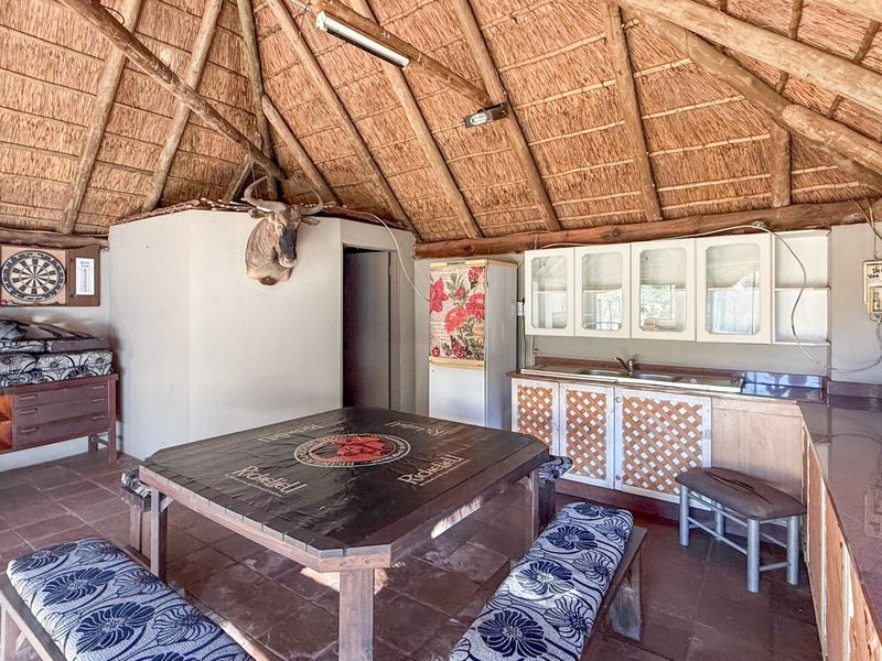 A rustic covered patio area featuring a traditional thatched roof, exposed timber rafters, built-in cabinetry with a sink, and tiled flooring.