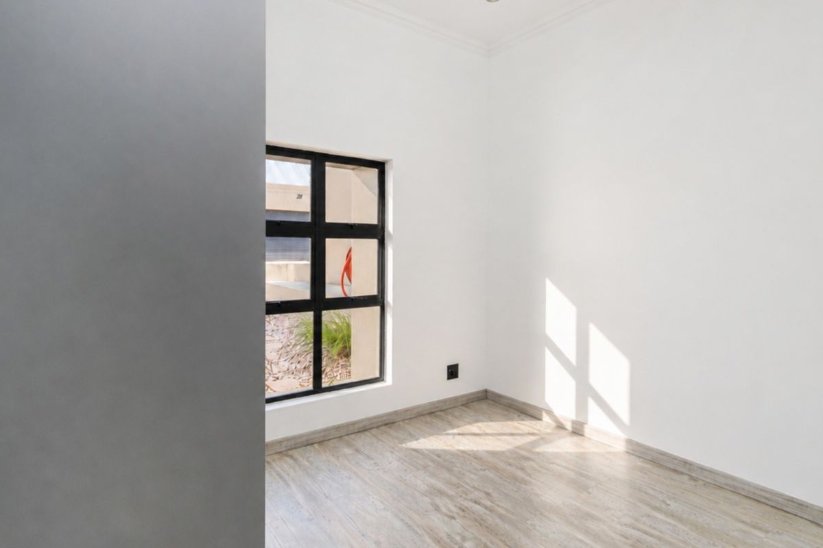 A bright, empty bedroom featuring modern wood-look laminate flooring, clean white walls, and a black-framed window allowing for natural light.