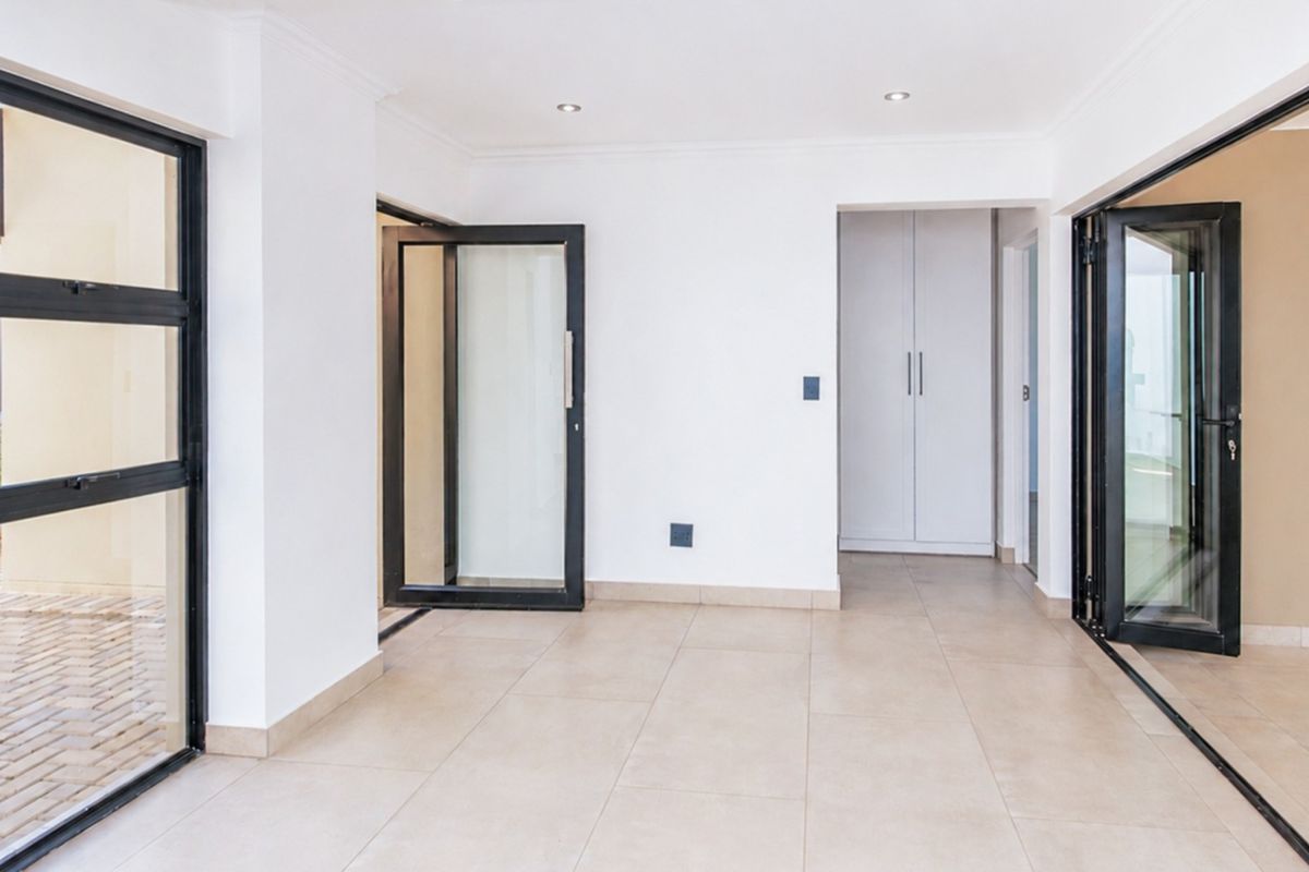 A bright, empty living area featuring neutral tiled flooring, recessed lighting, and modern black aluminium framed glass doors leading to the exterior.