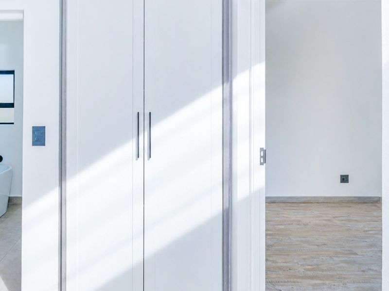 A bright, modern hallway featuring clean white built-in cupboards, light wood-look laminate flooring, and neutral finishes connecting to adjacent rooms.