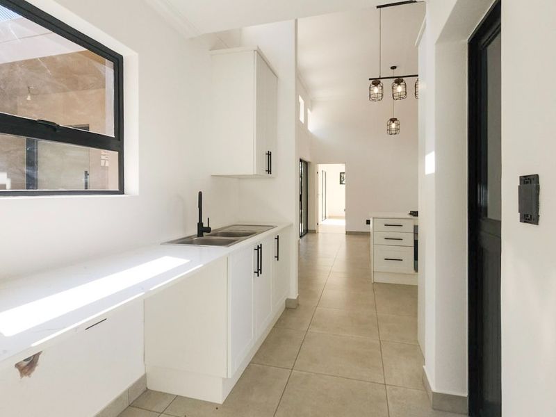 Modern kitchen featuring white cabinetry, stone countertops, double sink, and tiled flooring, leading into an open-plan living area with contemporary pendant lighting.