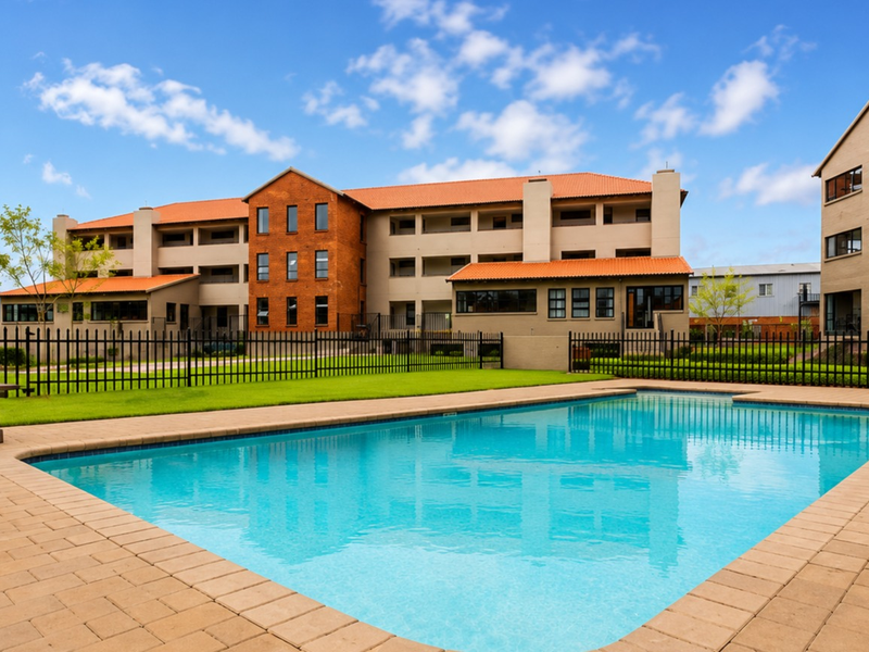 A well-maintained communal swimming pool area featuring paved decking, metal fencing, and views of the multi-story apartment complex with tiled roofing.
