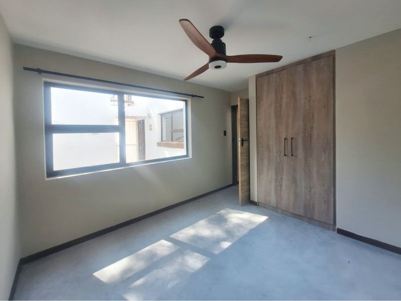 A bright, empty bedroom featuring a modern ceiling fan, built-in wooden wardrobe, and large aluminium-framed window.