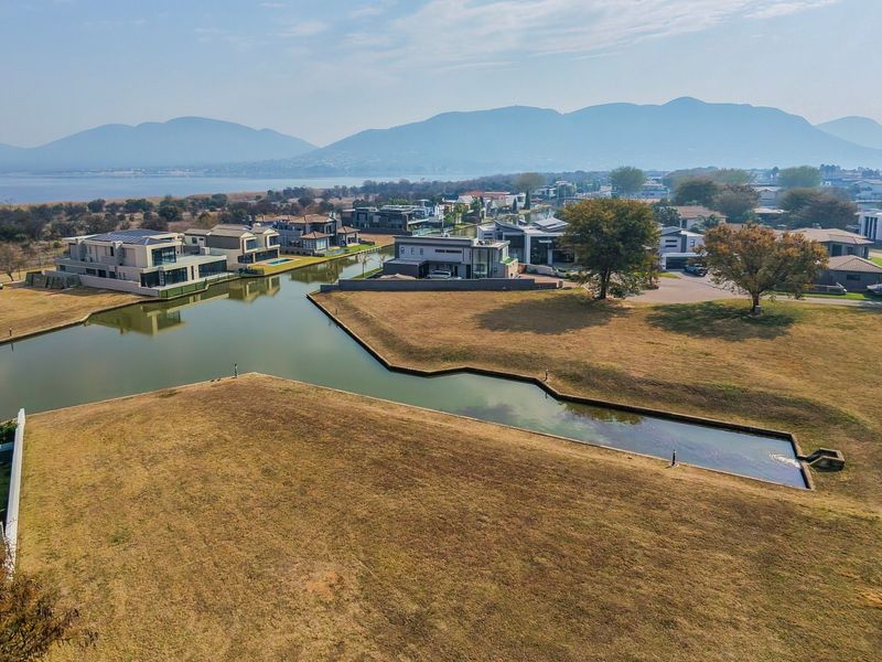 Aerial view of a residential estate featuring vacant land plots adjacent to a man-made canal with mountain and water views in the background.