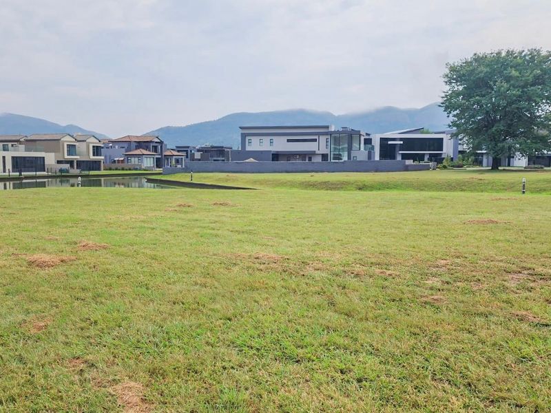 A scenic view of a vacant residential plot featuring a well-maintained lawn, a small water feature, and a backdrop of mountains and modern homes.