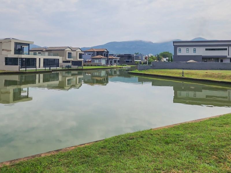 A scenic view of a residential canal with manicured lawns and modern homes in the background, set against a mountain landscape.