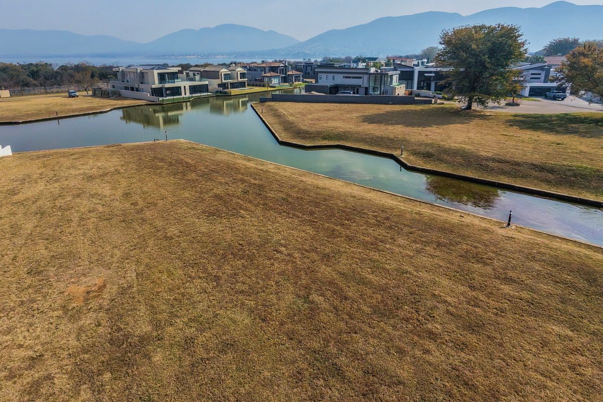 An elevated aerial view of a vacant residential plot situated along a canal, featuring scenic water views and surrounding estate homes.