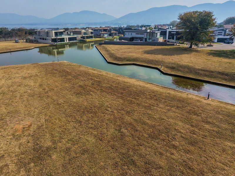 An elevated aerial view of a vacant residential plot situated along a canal, featuring scenic water views and surrounding estate homes.