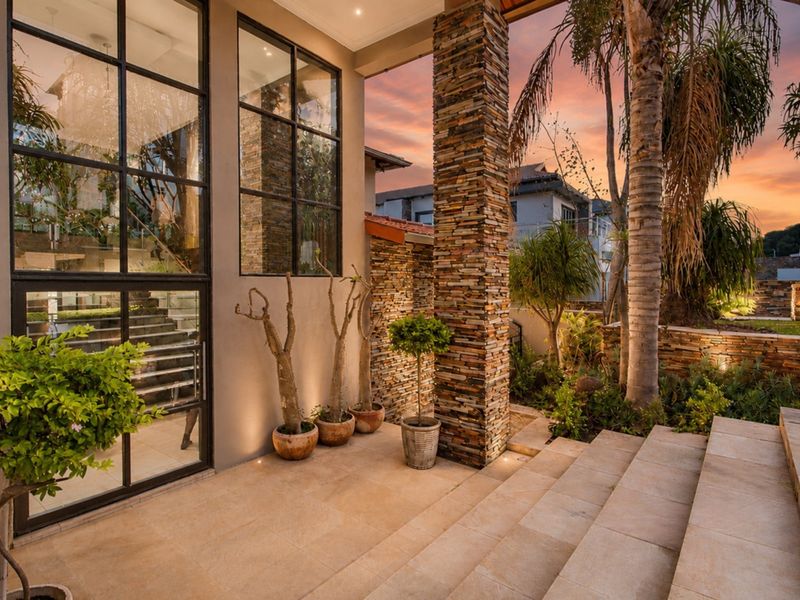 A beautifully designed covered patio featuring stone-clad pillars, large-format tiling, and expansive black steel-framed windows overlooking a landscaped garden area.