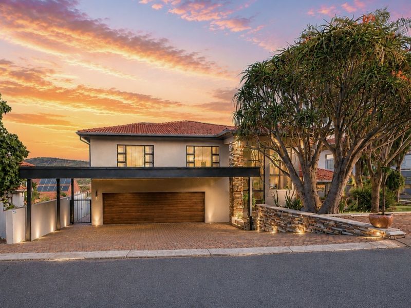 Front elevation of a modern house featuring a double garage, paved driveway, stone-clad retaining wall, and integrated exterior lighting at sunset.