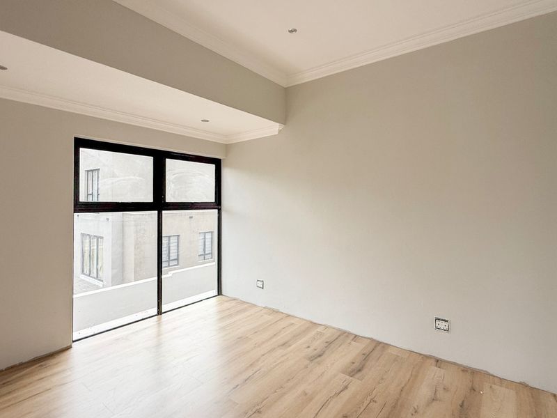 A bright, empty bedroom featuring modern laminate flooring, recessed downlighting, and large black-framed windows providing natural light.