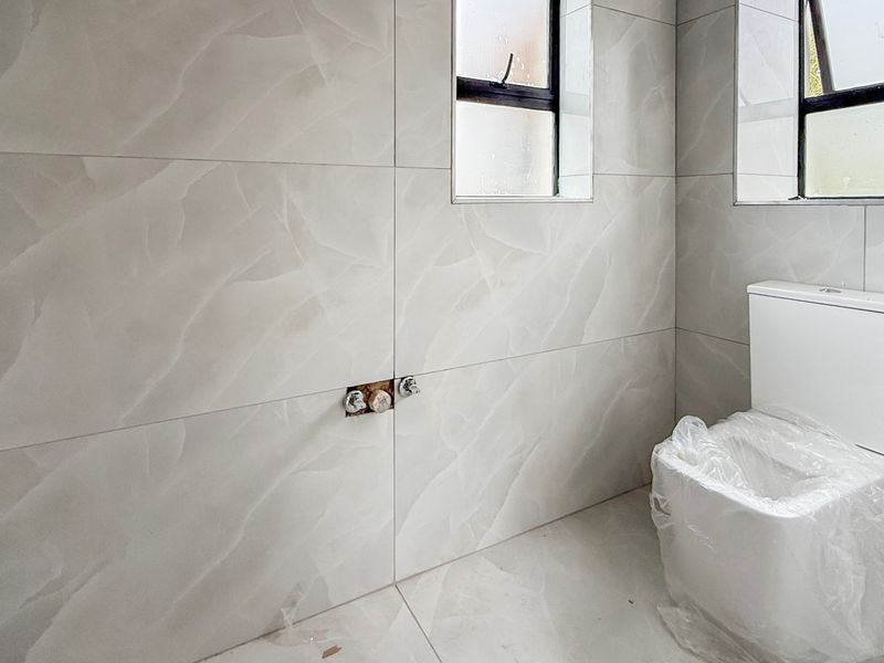 A modern bathroom featuring large-format light grey marble-effect tiles, a new dual-flush toilet, and an unfinished plumbing connection point on the wall.