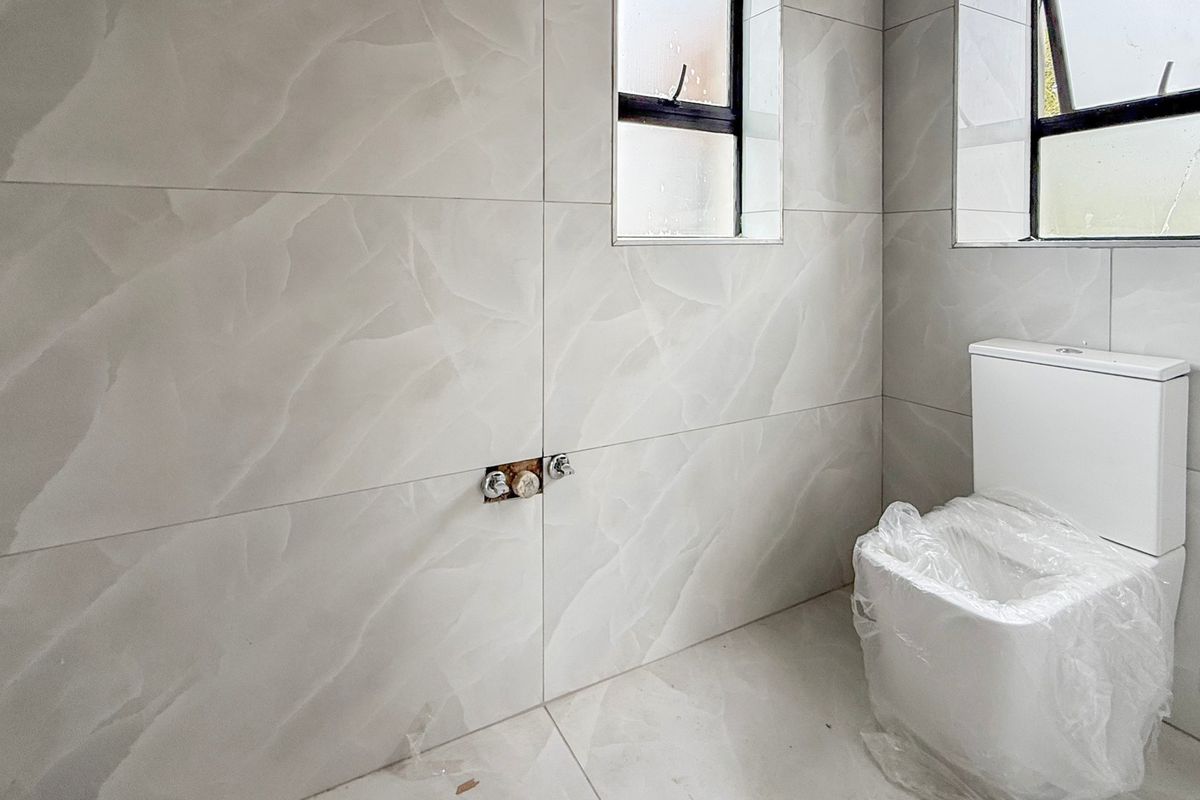 A modern bathroom featuring large-format light grey marble-effect tiles, a new dual-flush toilet, and an unfinished plumbing connection point on the wall.