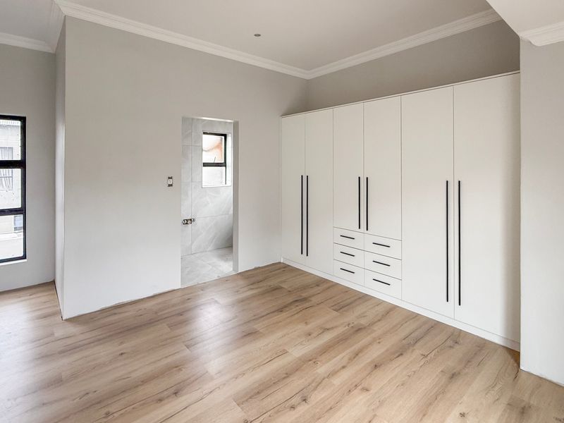 A bright, modern bedroom featuring light wood-look laminate flooring, extensive built-in cabinetry with sleek handles, and a neutral color palette.