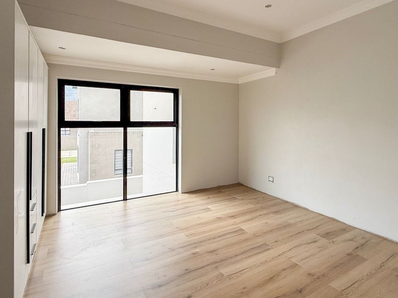 A bright, modern bedroom featuring light wood-look laminate flooring, built-in cupboards, and large aluminium-framed windows.
