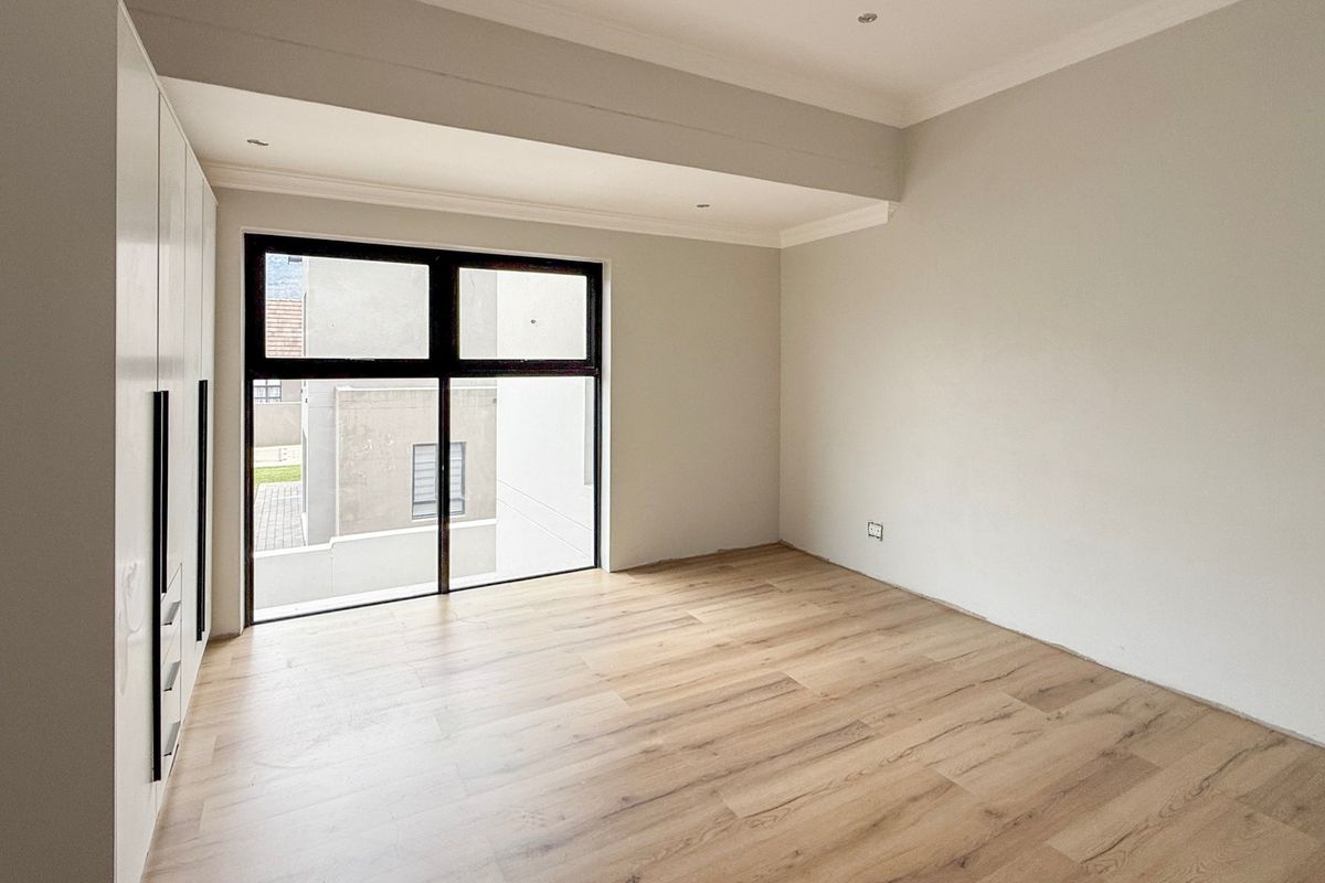 A bright, modern bedroom featuring light wood-look laminate flooring, built-in cupboards, and large aluminium-framed windows.