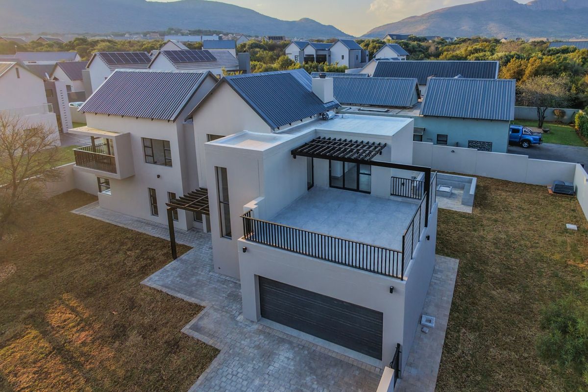 Aerial view of a modern double-story residential property featuring a metal roof, balcony, garage, and paved outdoor areas within a secure estate.