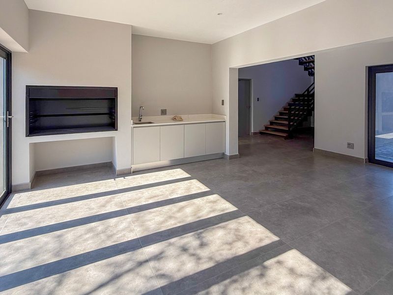 Spacious open-plan living area featuring a built-in braai, kitchenette with sink, modern tiled flooring, and large sliding doors leading to the outdoor patio.
