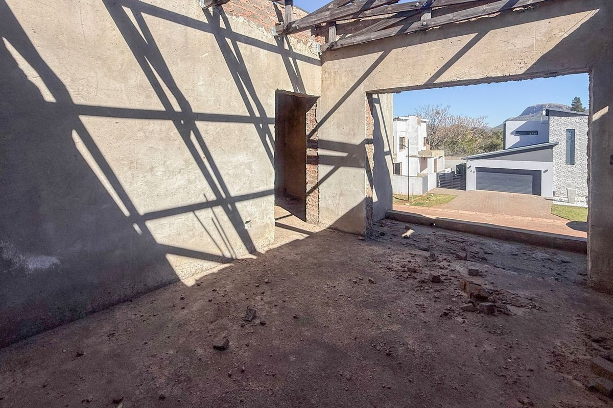 An unfinished interior space under construction featuring exposed roof trusses, plastered masonry walls, and a large opening overlooking the neighborhood.