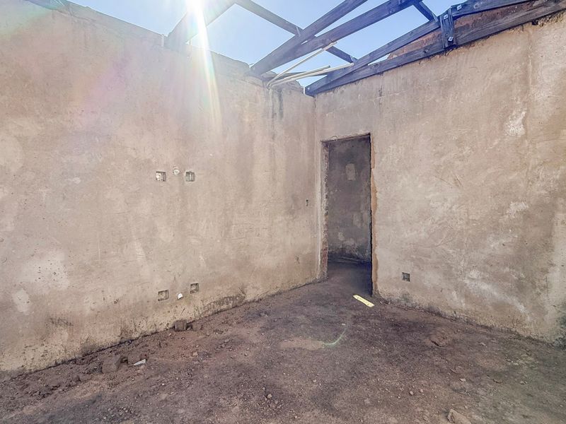 A shell-condition interior space featuring exposed roof rafters, unfinished masonry walls, and an earthen floor, requiring significant structural renovation.