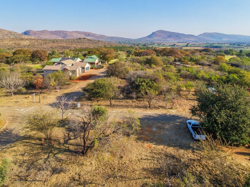 An aerial view of a vacant residential plot within a scenic estate, featuring established trees, natural landscape, and access to paved estate roads.