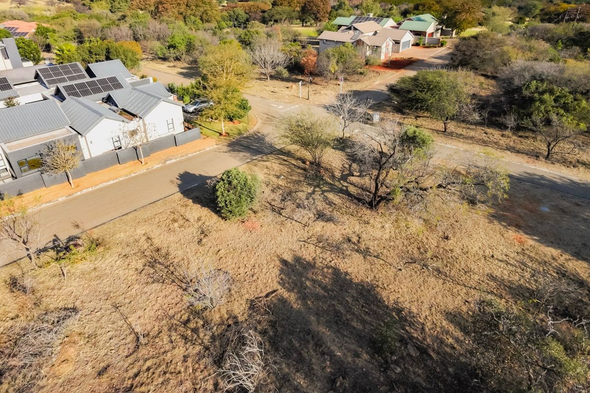 An aerial view of a vacant residential plot situated in a developed estate, featuring natural vegetation and adjacent paved road access.