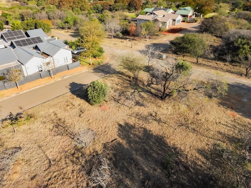 An aerial view of a vacant residential plot situated in a developed estate, featuring natural vegetation and adjacent paved road access.